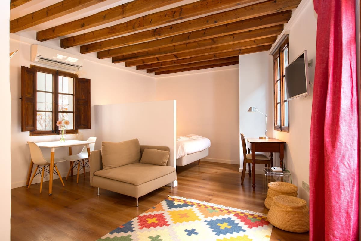 A cozy room with a wooden ceiling, a bed, a sofa, a dining table with chairs, and a TV on the wall.
