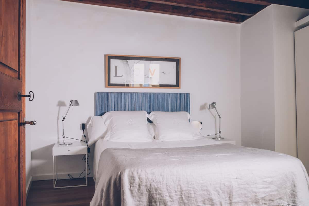 A bedroom with a bed, two bedside tables, a nightstand, a framed artwork on the wall, and a wooden door.