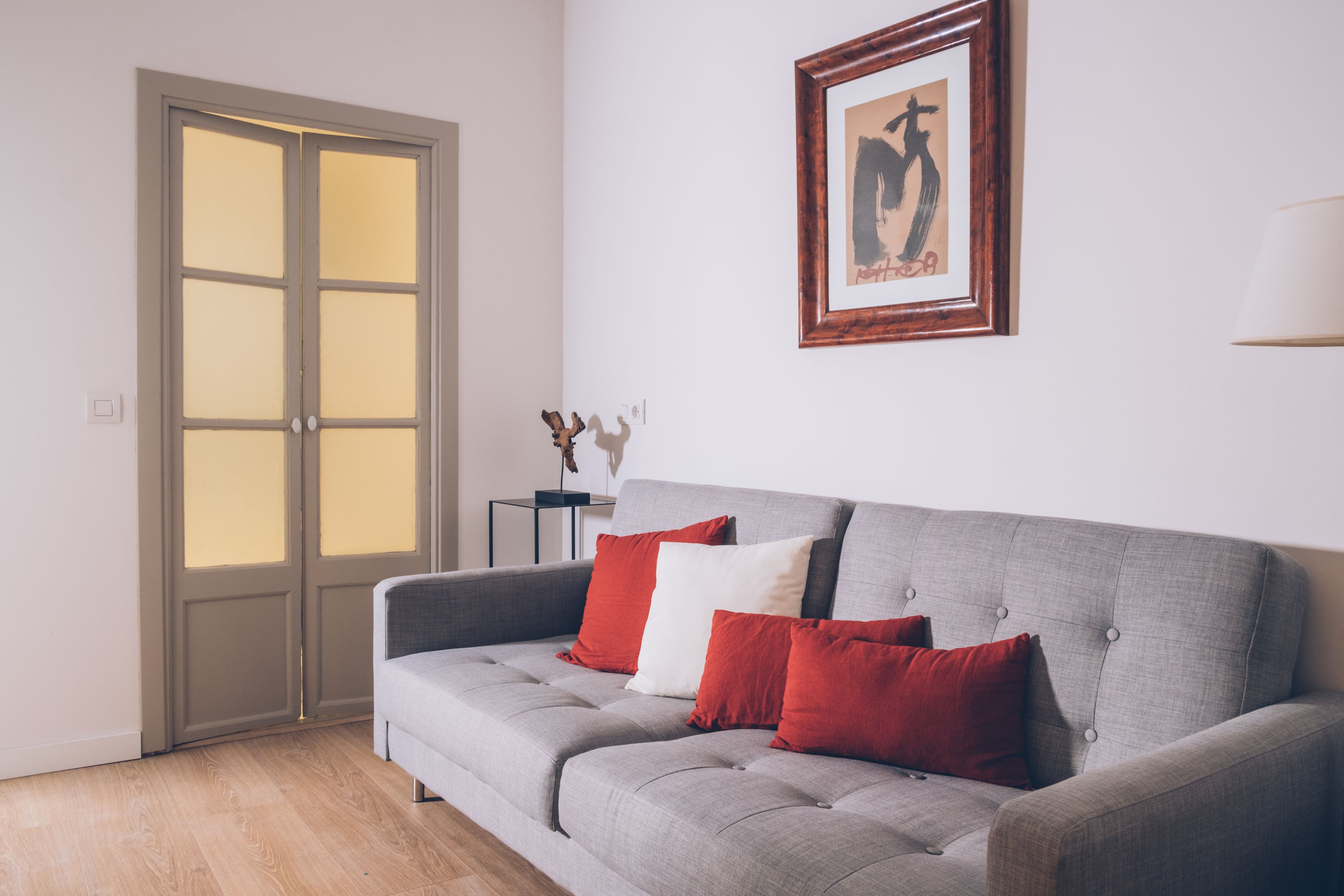 Una sala de estar moderna con un sofá gris, cojines rojos y blancos, una obra de arte enmarcada en la pared y piso de madera.