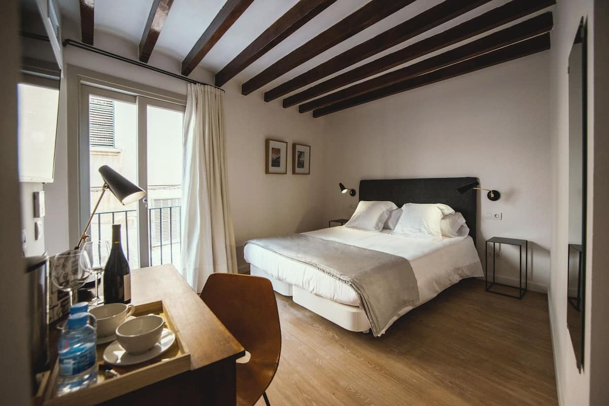 A bedroom with a bed, bedside tables, a desk with a chair, a window with curtains, and wooden beams on the ceiling.
