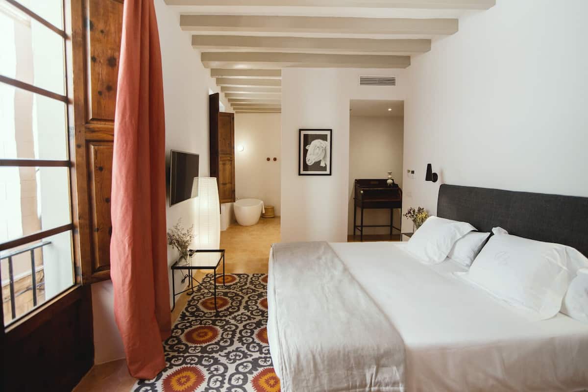 A hotel room with a bed, a desk, a chair, a window with curtains, a bathroom, and a hallway.
