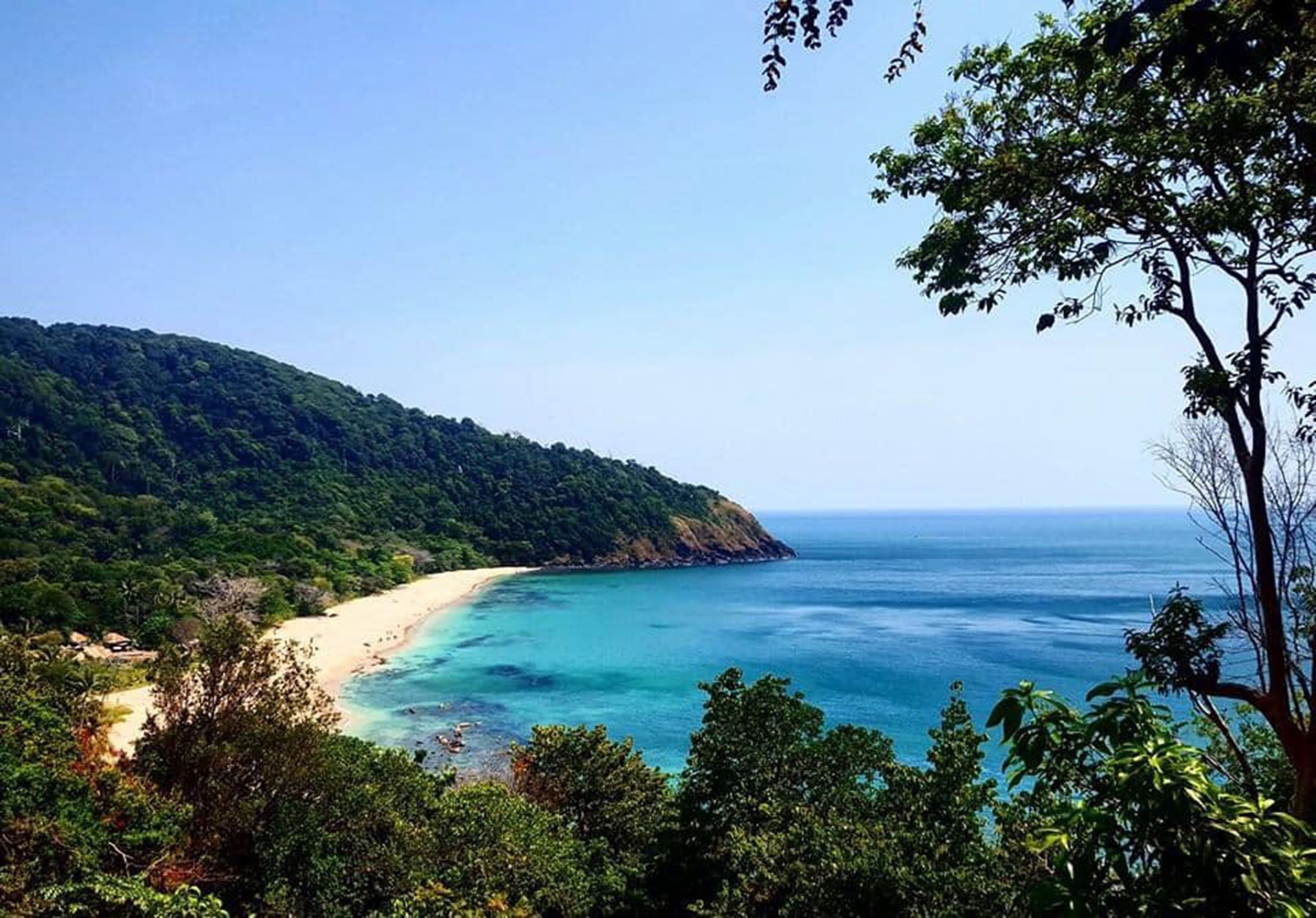 A coastal landscape with a beach, forested hills, and clear blue waters.