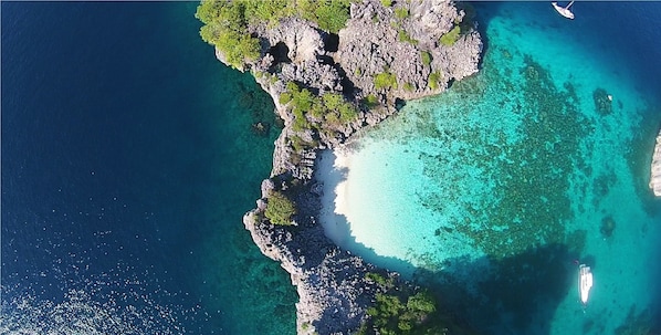 Vista aérea de una zona costera con aguas turquesas cristalinas, terreno rocoso y un pequeño bote anclado cerca de la orilla.