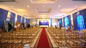 A banquet hall with rows of chairs, a red carpet, chandeliers, and a stage with a large screen. - Trang An Hotel (Hoa Lu)