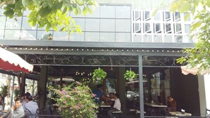An outdoor cafe with tables and chairs, surrounded by greenery and a building with large windows in the background.