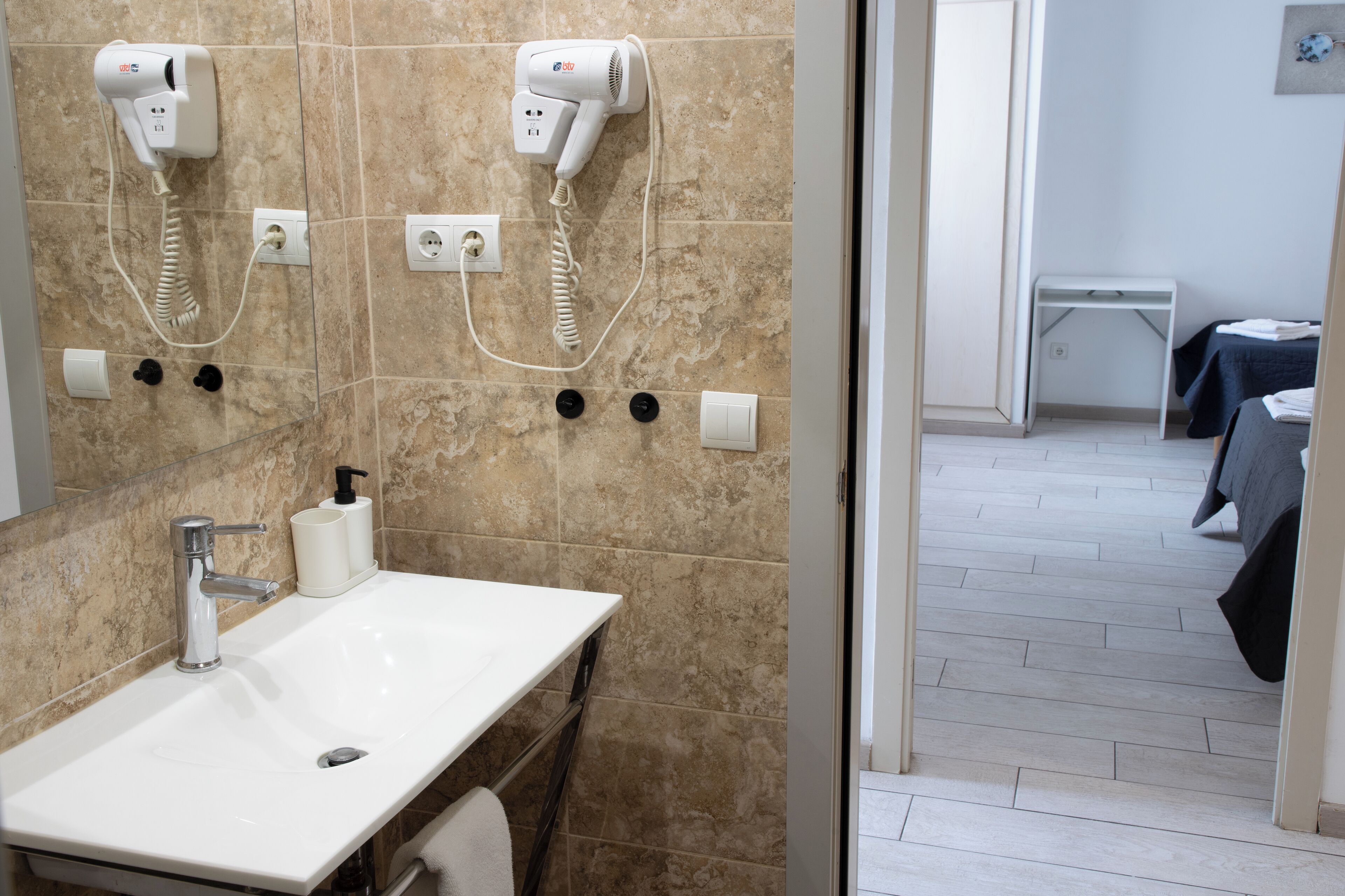 A bathroom with a sink, two hair dryers mounted on the wall, and a small table with a towel.