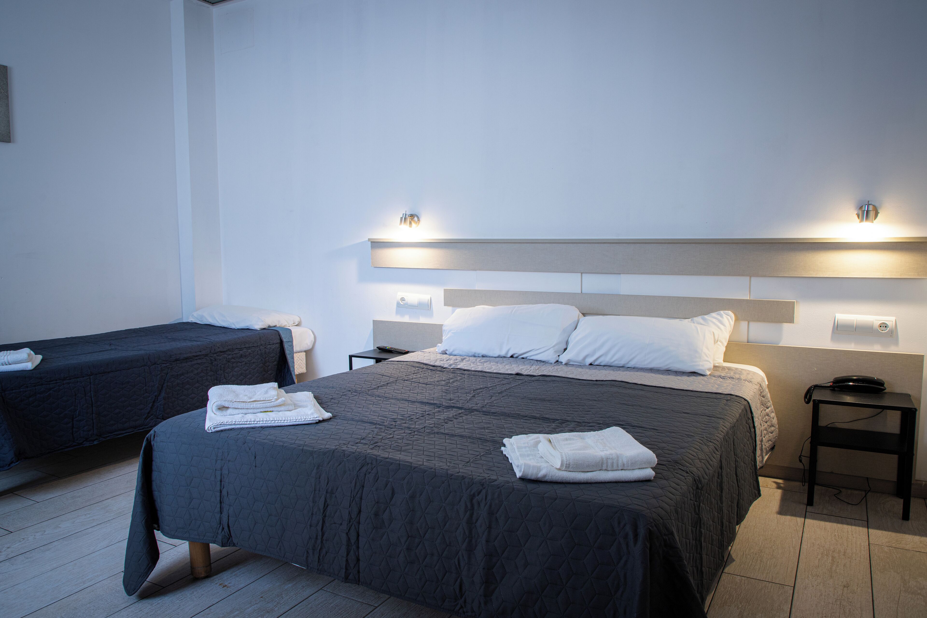 Ein Hotelzimmer mit zwei Betten, jeweils mit einer dunkel gesteppten Tagesdecke und weißen Kissen. An beiden Betten befinden sich Nachttische mit Lampen und ein an der Wand montiertes Telefon.