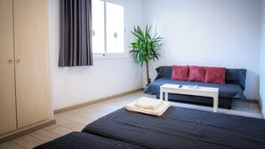 Ein Zimmer mit zwei Betten, einem Schlafsofa, einem kleinen Tisch, einer Pflanze und einem Fenster mit Vorhängen.