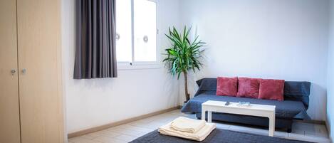 A room with two beds, a sofa, a small table, a plant, and a window with curtains.