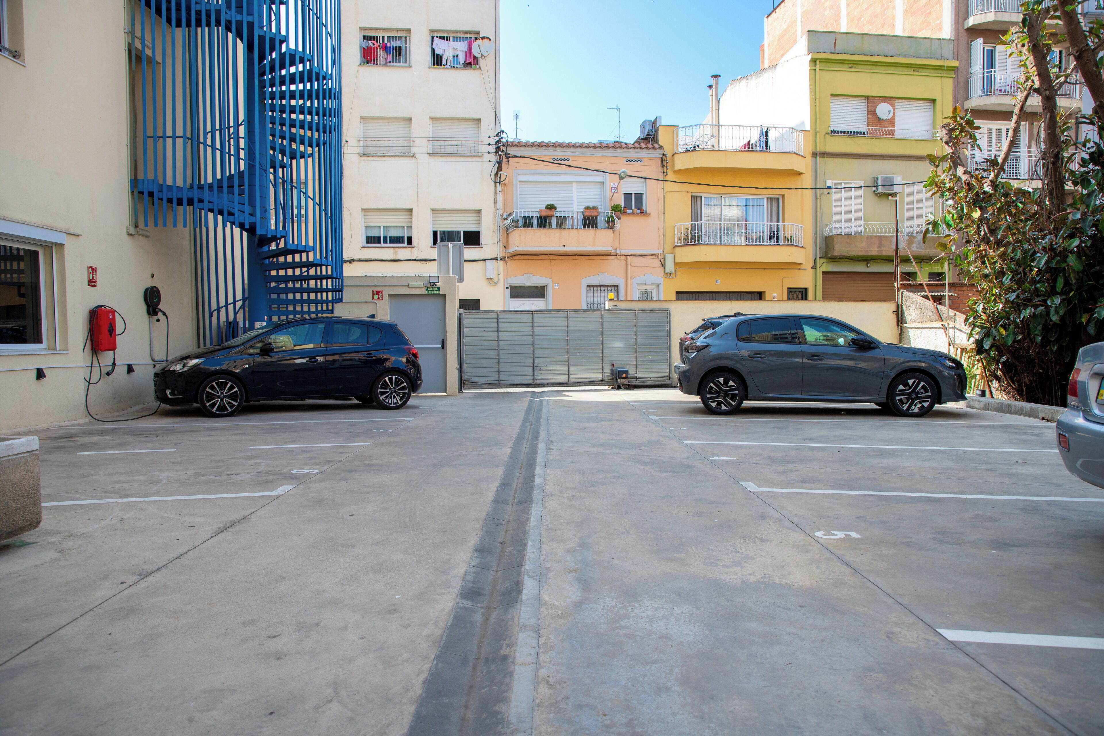Un parking où sont garées deux voitures devant un immeuble doté d’un escalier en colimaçon bleu et de balcons.