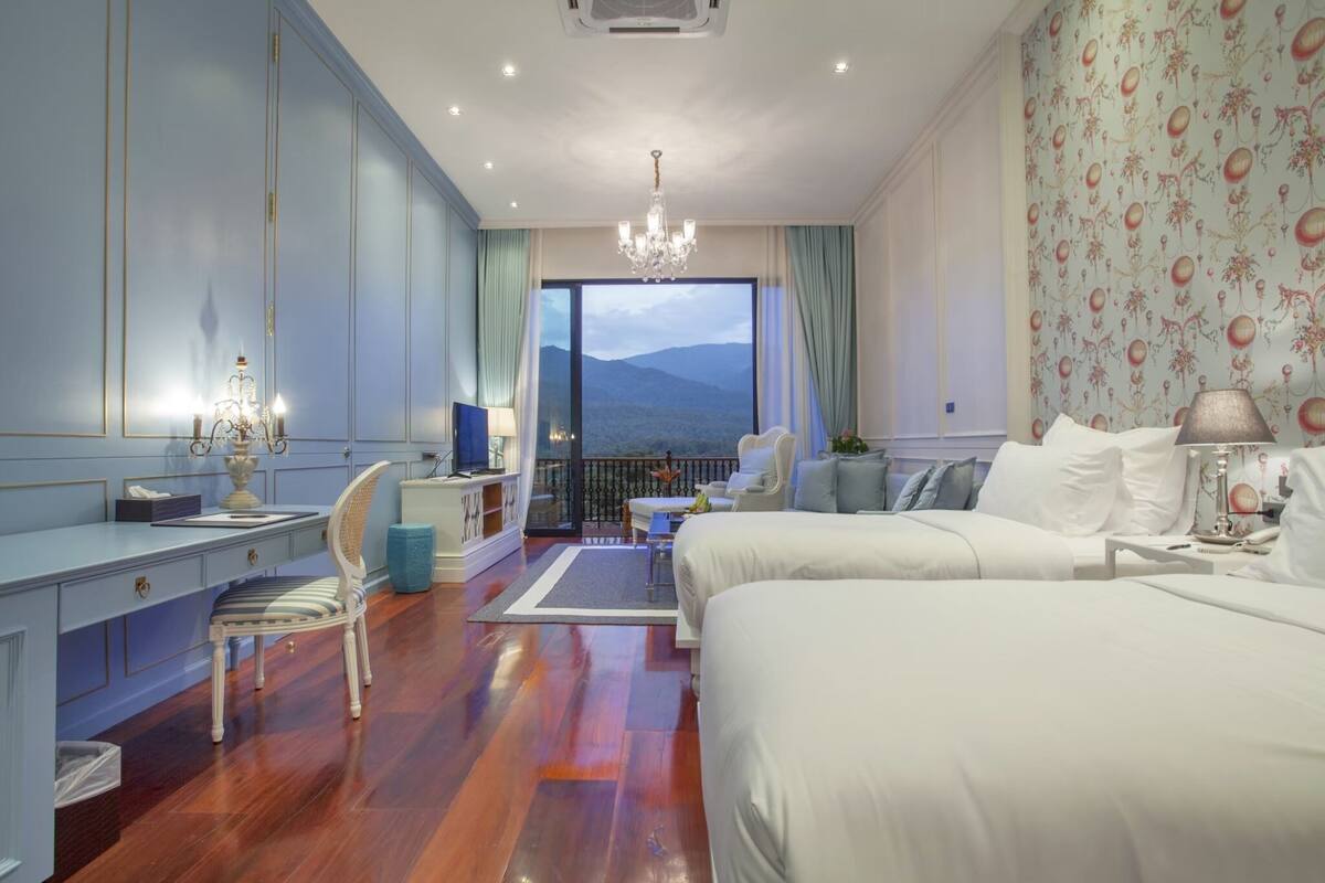 A hotel room with a large bed, a desk with a chair, a television, and a balcony with a view of the mountains.