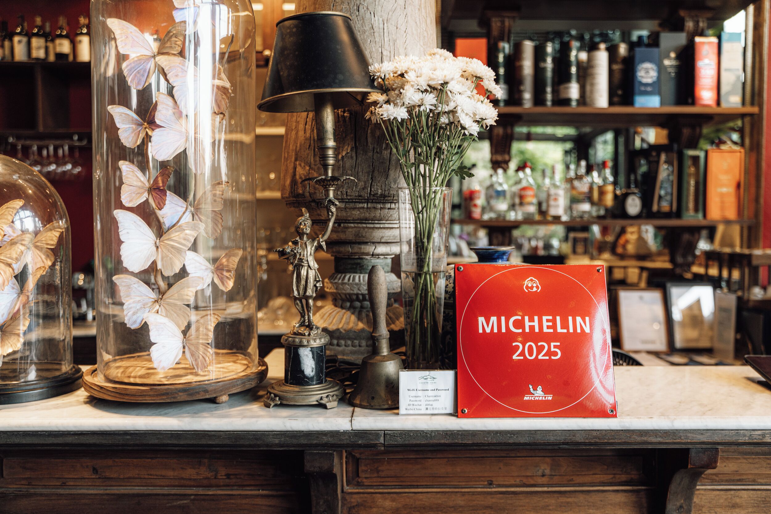 A display of decorative items including glass vases with butterflies, a vintage lamp, and a bouquet of flowers. A red sign with 'MICHELIN 2025' is also visible.