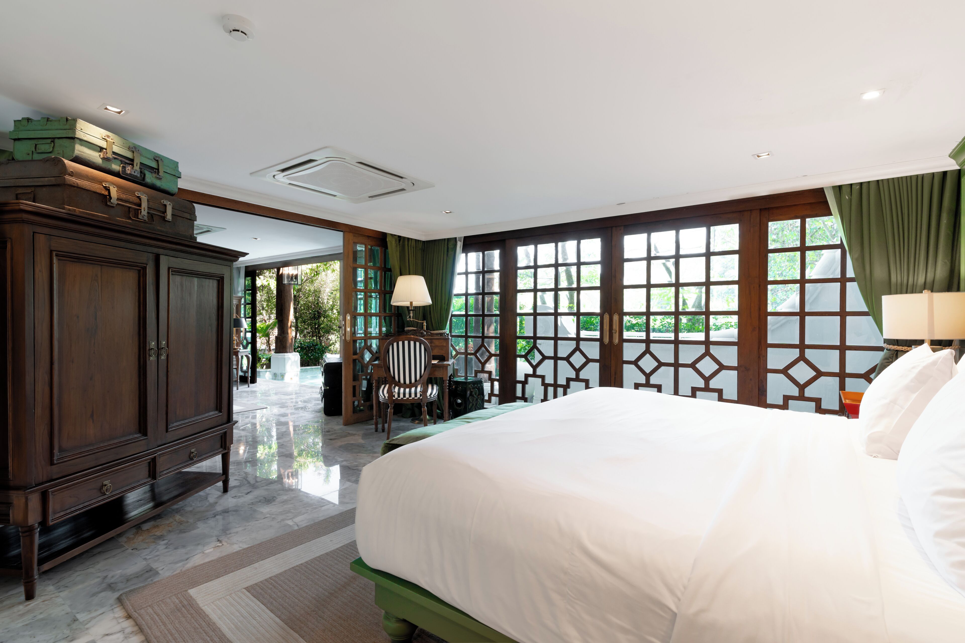 A spacious hotel room with a large bed, a wooden wardrobe, a desk with a chair, and a view through large windows.