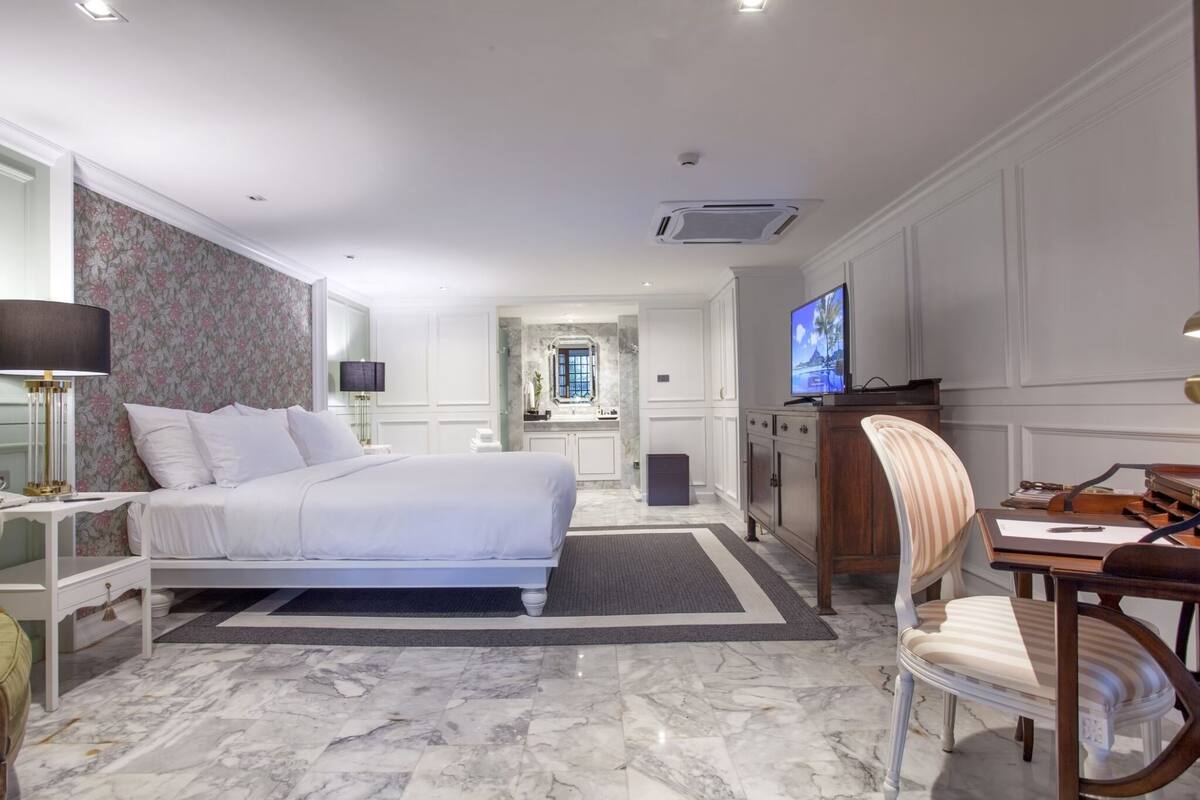 A hotel room with a large bed, a desk with a chair, a television, and a bathroom visible through an archway.