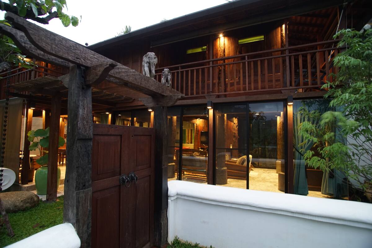 A wooden house with a balcony, a covered patio, and a well-lit interior.
