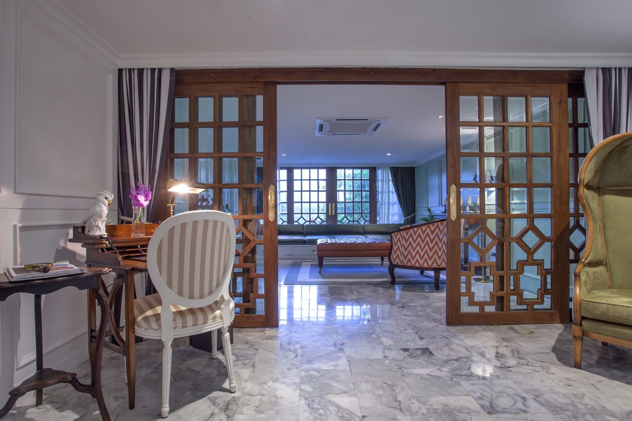 A room with a marble floor, a desk with a lamp, a chair, and a sofa. There are wooden doors with glass panels leading to another room.