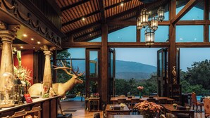A spacious dining area with a large wooden table, comfortable seating, and a view of the mountains through large windows. - Chantra Khiri Chalet Chiang Mai (Hang Dong)