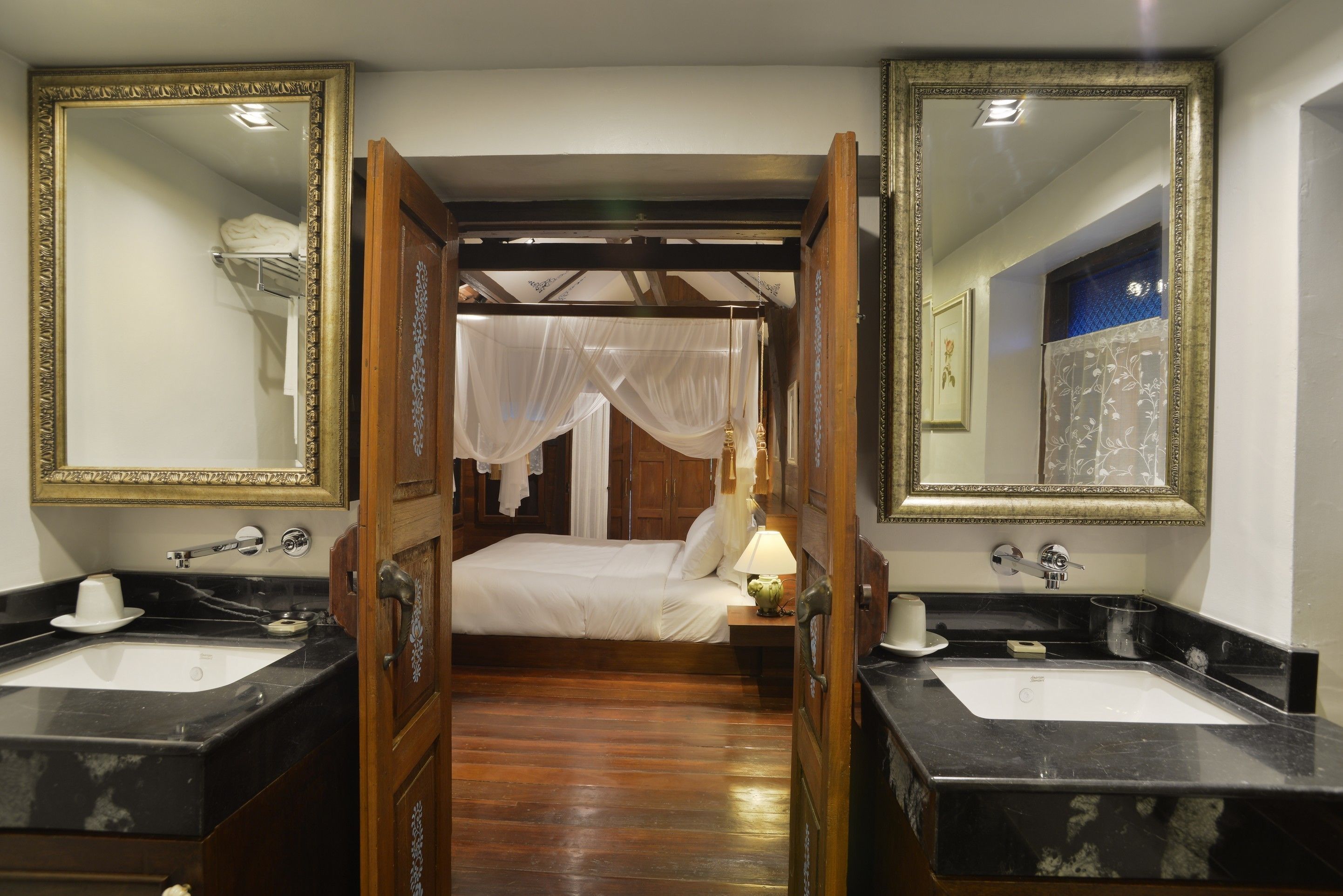 A bedroom with a bed, two bedside tables, a nightstand, a mirror, and a bathroom with a sink and mirror.