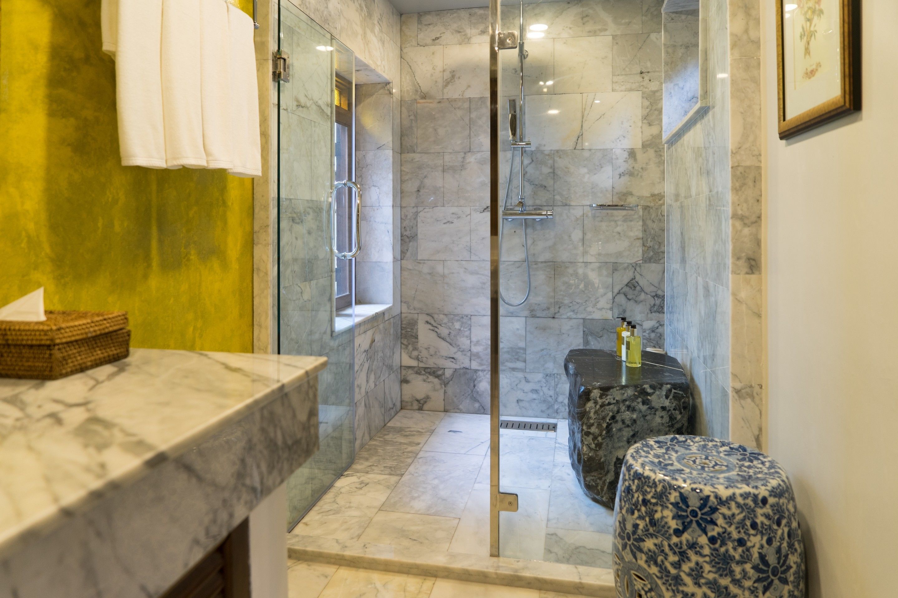 Un baño moderno con ducha de vidrio, paredes de mármol y un taburete pequeño con estampado floral.