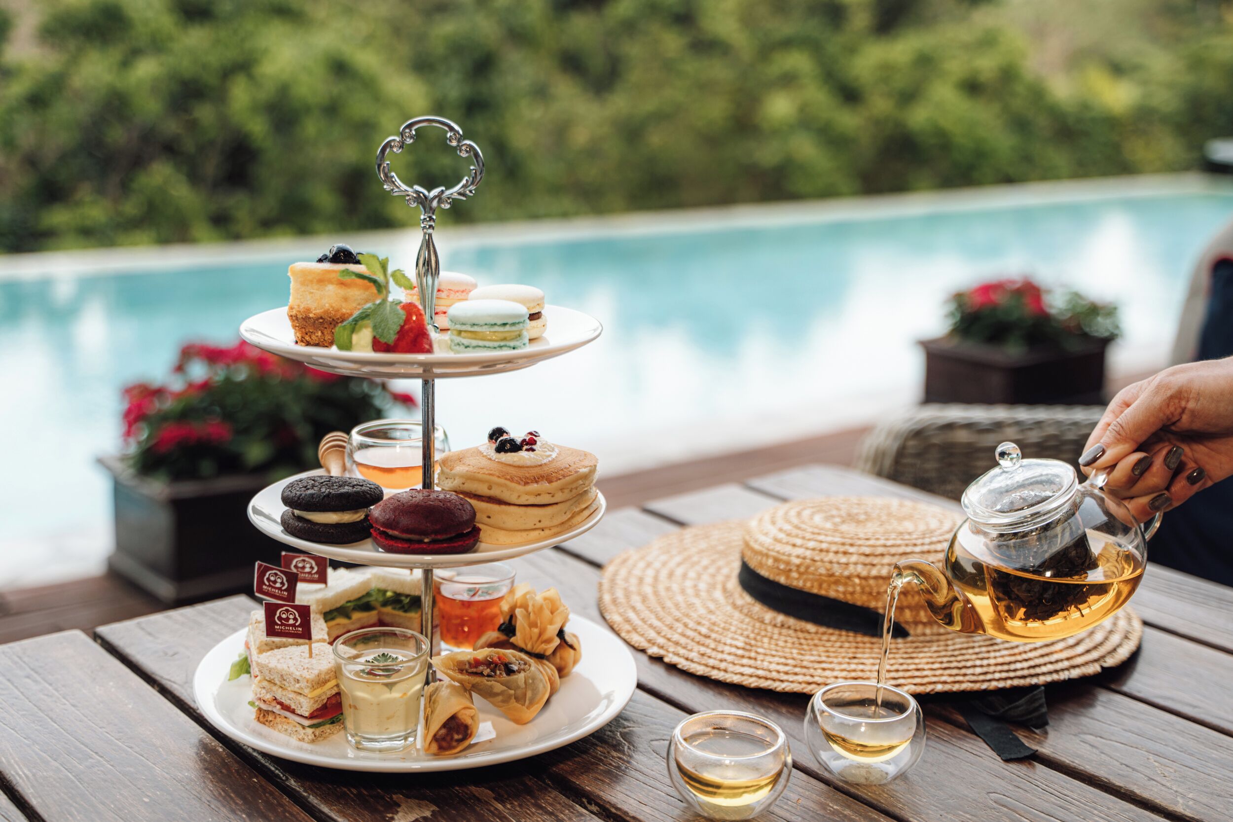 Ein mehrstöckiges Desserttablett mit Macarons, Kuchen und Sandwiches, eine Teekanne, aus der Tee ausgeschenkt wird, und im Hintergrund ein Schwimmbad.