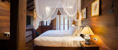 A wooden four-poster bed with white linens, a bedside table with a lamp and a vase, and a framed picture on the wall.