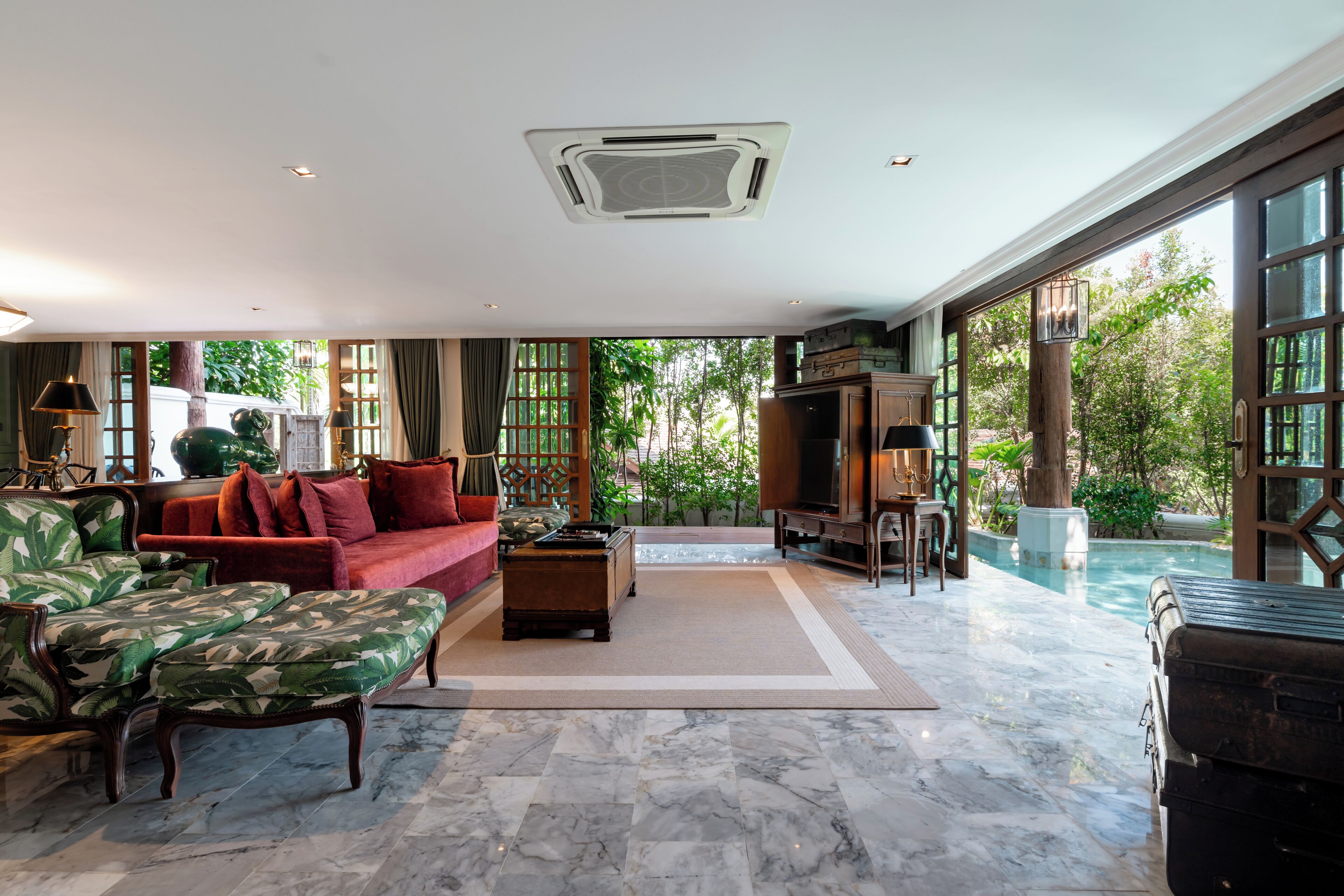 A spacious living room with a red sofa, two armchairs, a coffee table, and a TV. Large windows offer a view of a swimming pool and greenery.