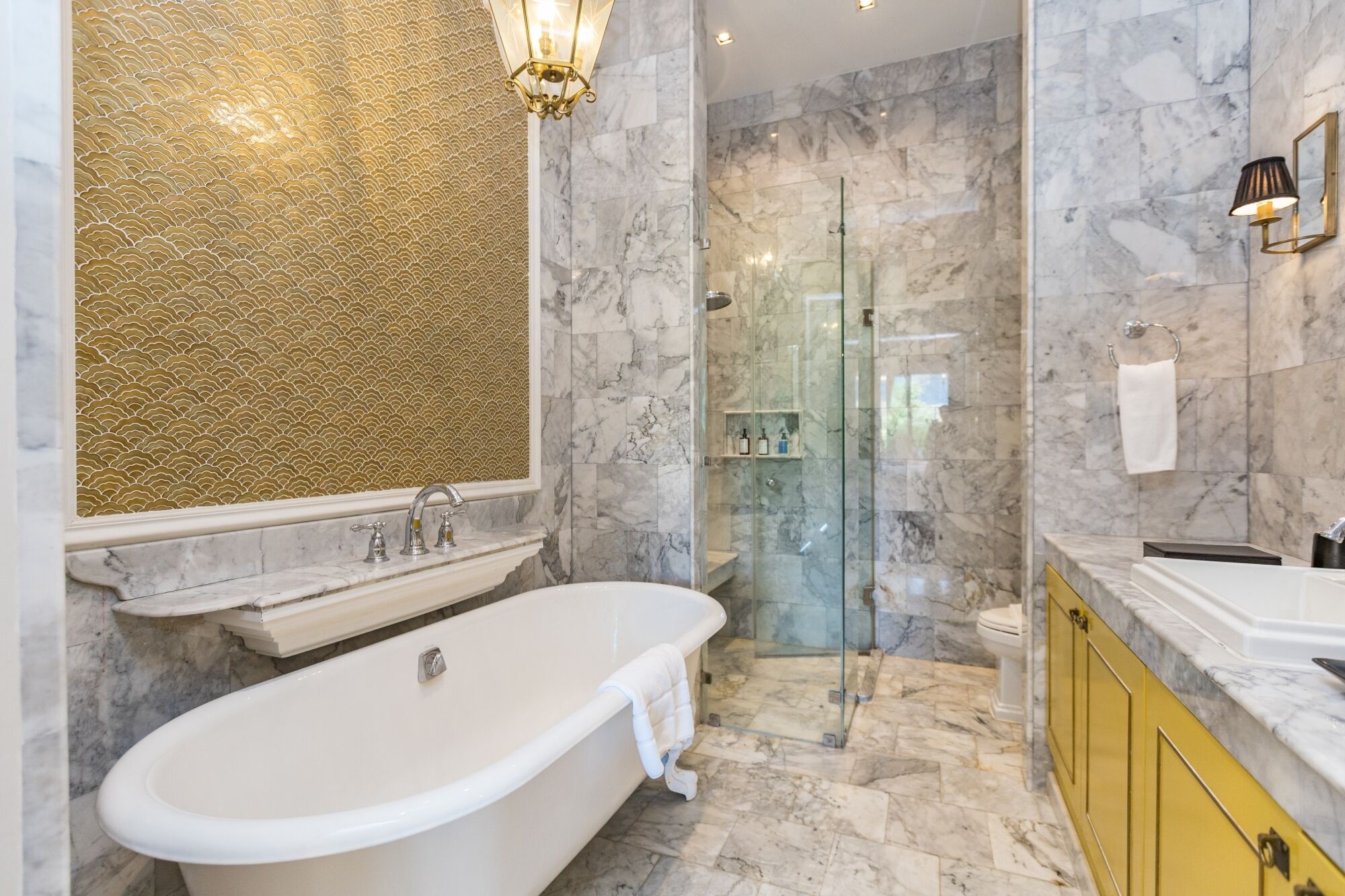 A bathroom with a freestanding bathtub, a glass-enclosed shower, marble walls, and a yellow vanity.