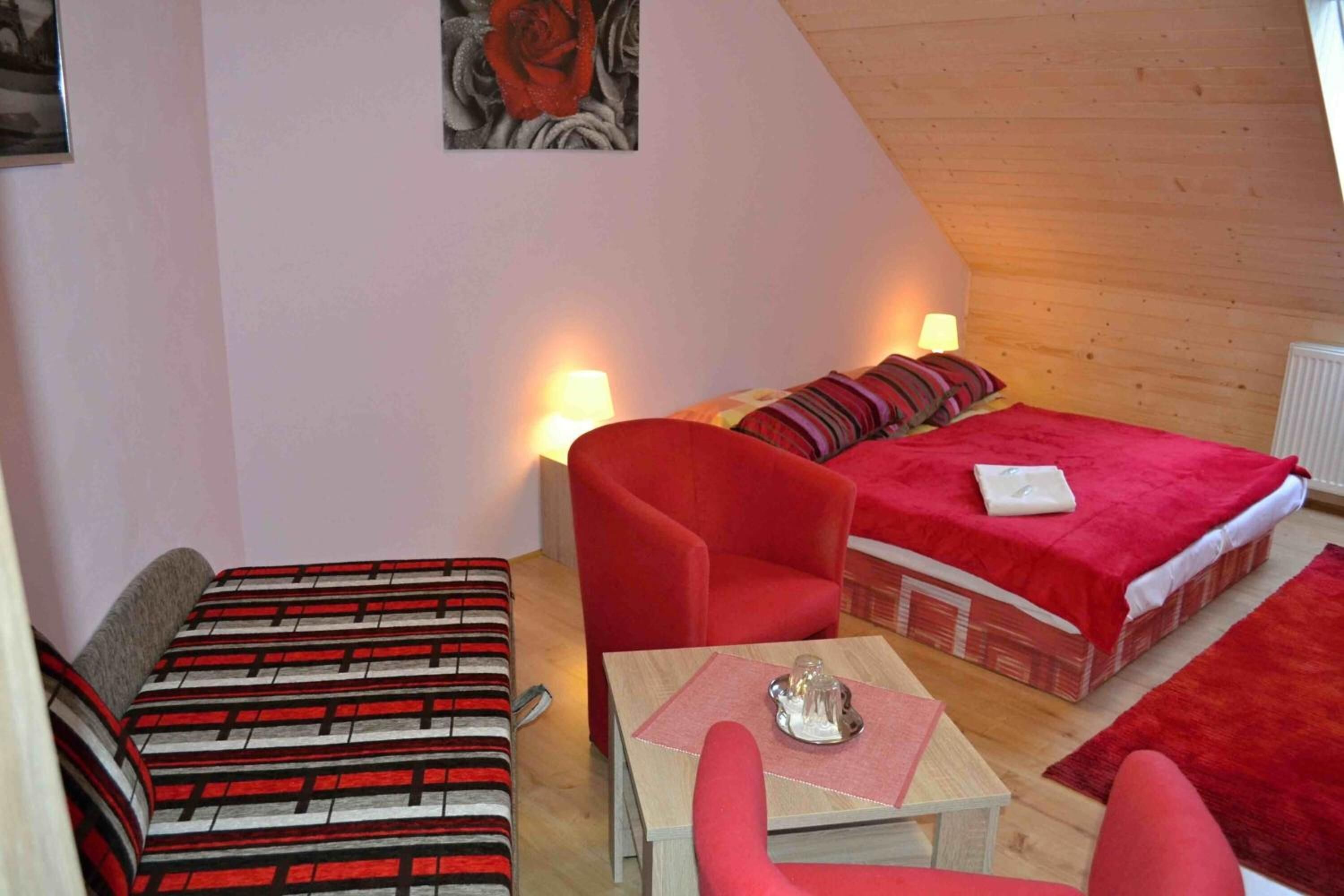 Quarto aconchegante com uma cama, duas cadeiras vermelhas, uma mesa pequena com abajur e um quadro na parede.