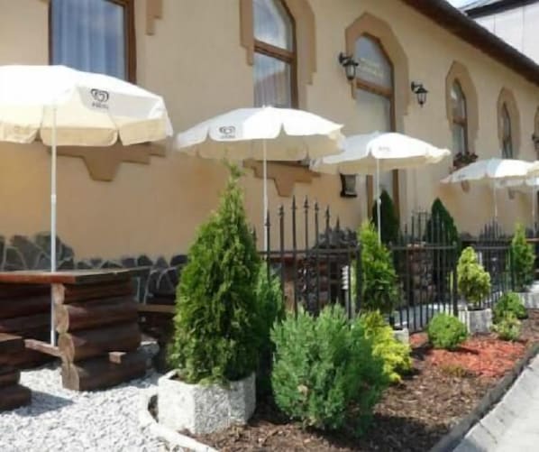 An outdoor seating area with tables and umbrellas, surrounded by decorative shrubs and a metal fence.