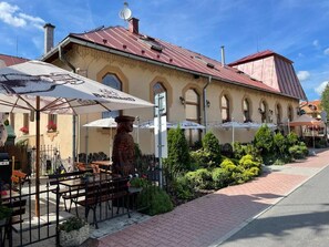 Un café en plein air avec des tables et des chaises, des parasols, et un bâtiment avec un toit rouge en arrière-plan.