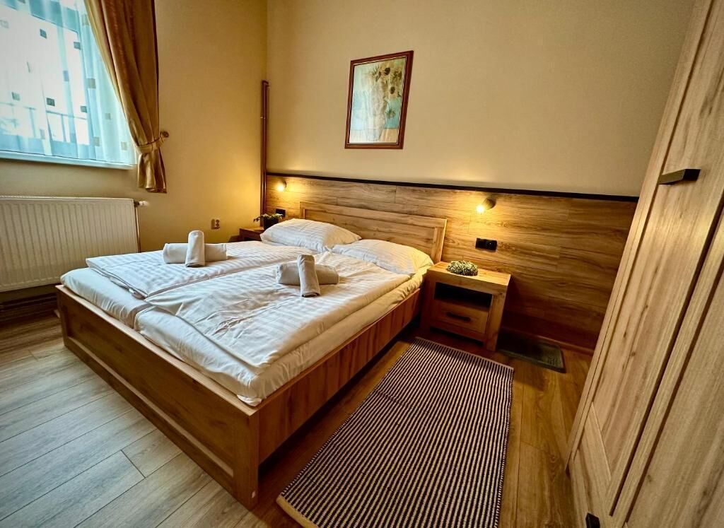 Una habitación de hotel con una cama de madera, una mesita de noche, un cuadro enmarcado en la pared y una ventana con cortinas.