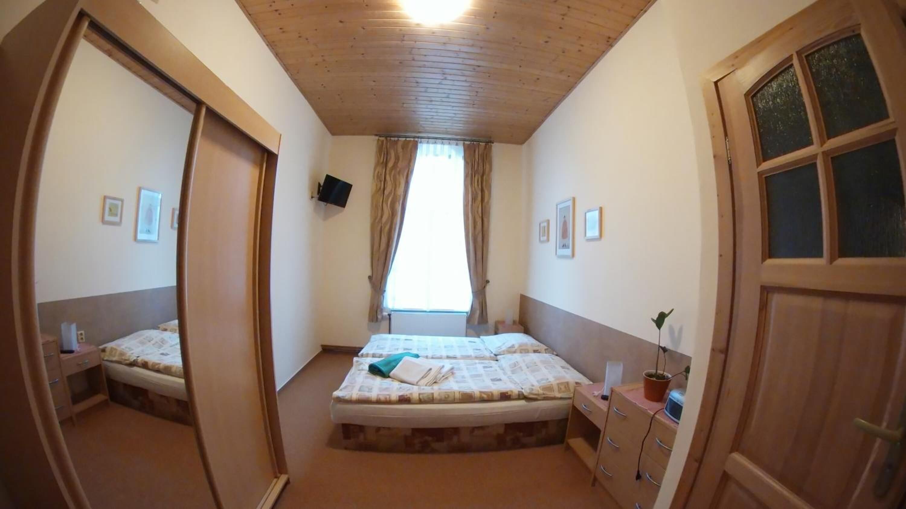 A bedroom with a wooden ceiling, a bed with a white mattress and a blue blanket, a nightstand with a lamp, and a window with curtains.