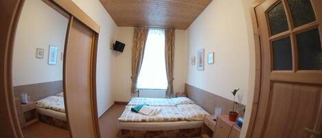 A bedroom with a wooden ceiling, a bed with a white mattress and a blue blanket, a nightstand with a lamp, and a window with curtains.