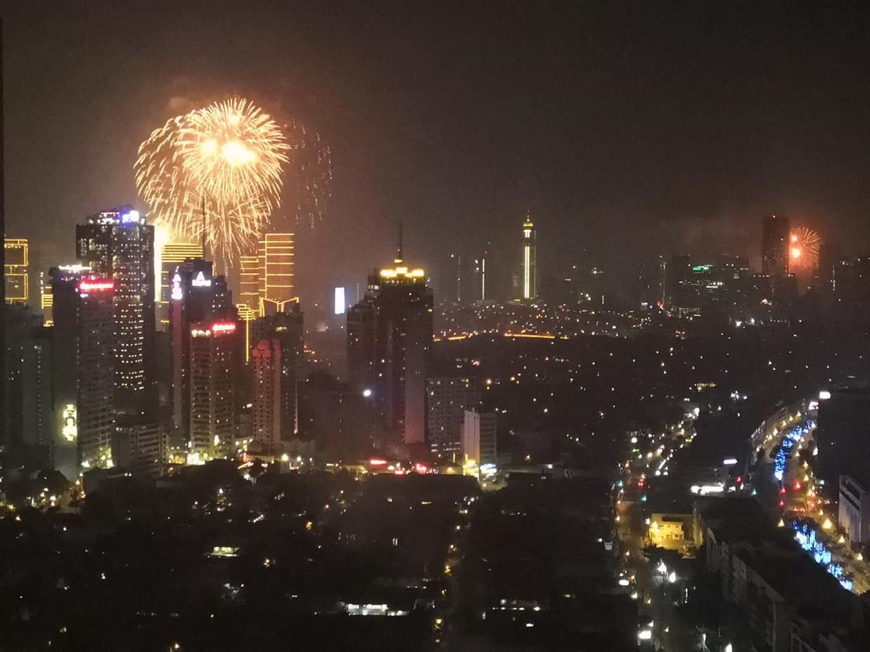 Une vue nocturne d’une ville, avec des bâtiments illuminés et des feux d’artifice dans le ciel.