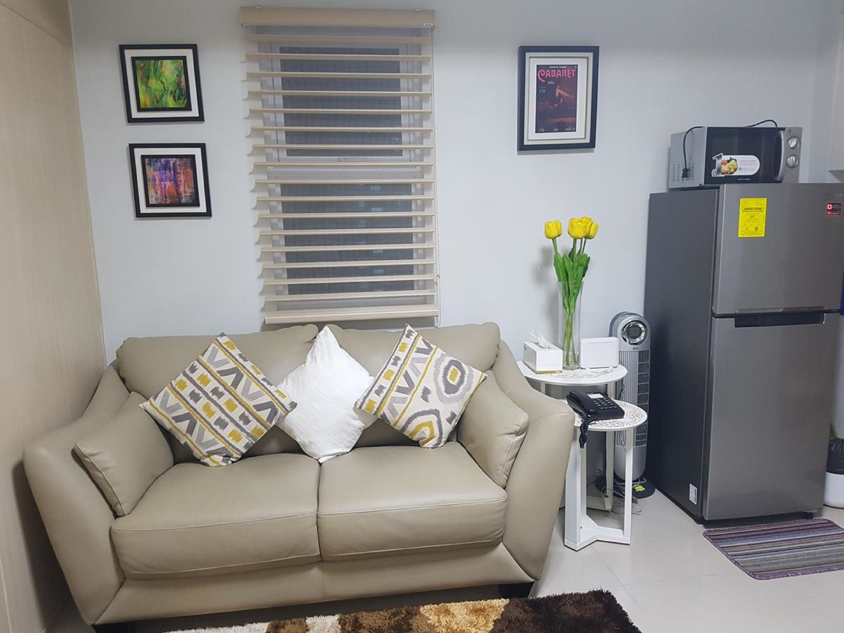A living room with a beige sofa, two pillows, a small side table with a vase of yellow flowers, a refrigerator, and a microwave.