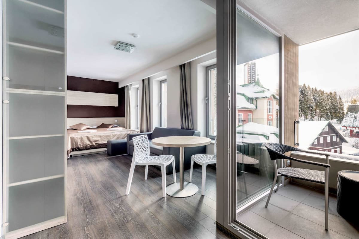 A modern hotel room with a balcony, a dining area with white chairs, and a view of a snowy town.