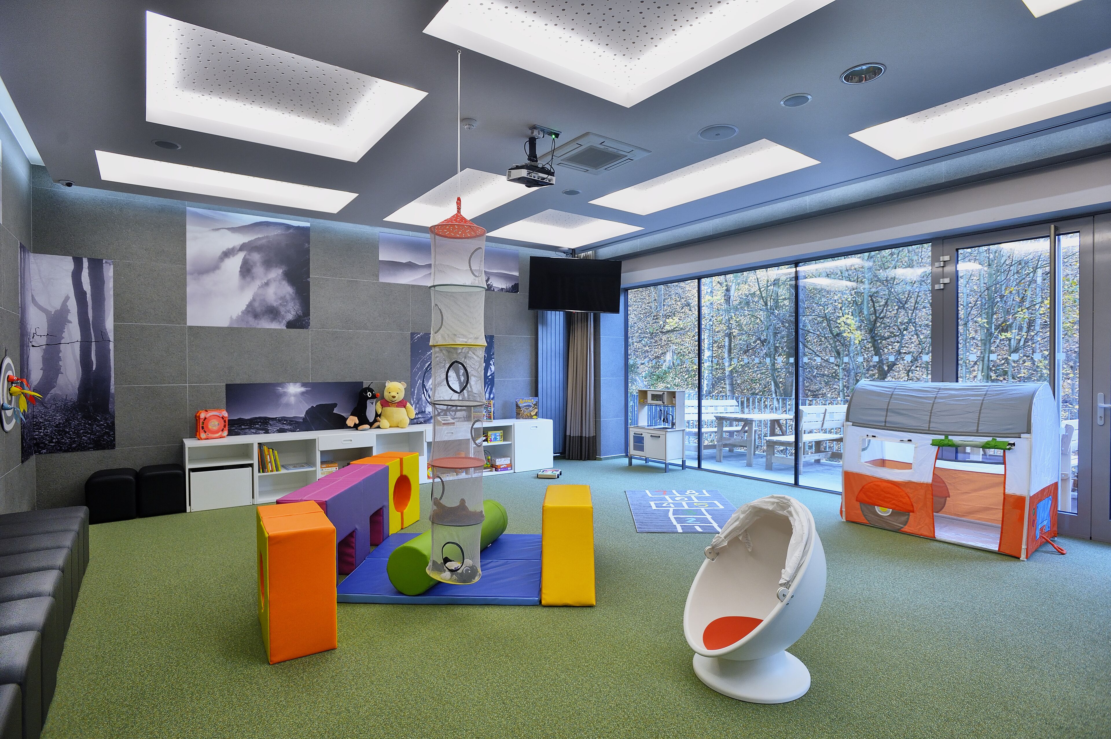 Une salle de jeux moderne, dotée de structures colorées, d’un téléviseur fixé au mur et de grandes fenêtres offrant une vue sur l’extérieur.