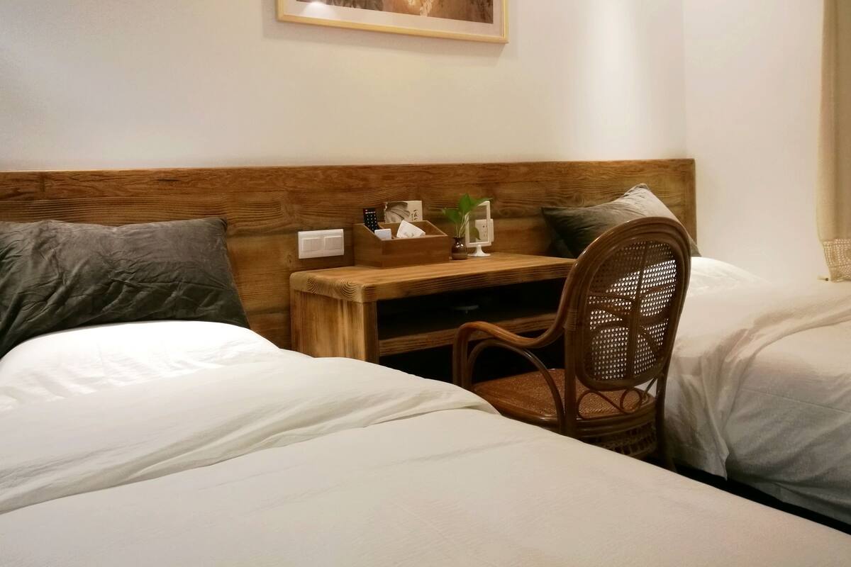 Quarto de hotel com duas camas, cabeceira de madeira, pequena escrivaninha com uma planta e um quadro na parede.