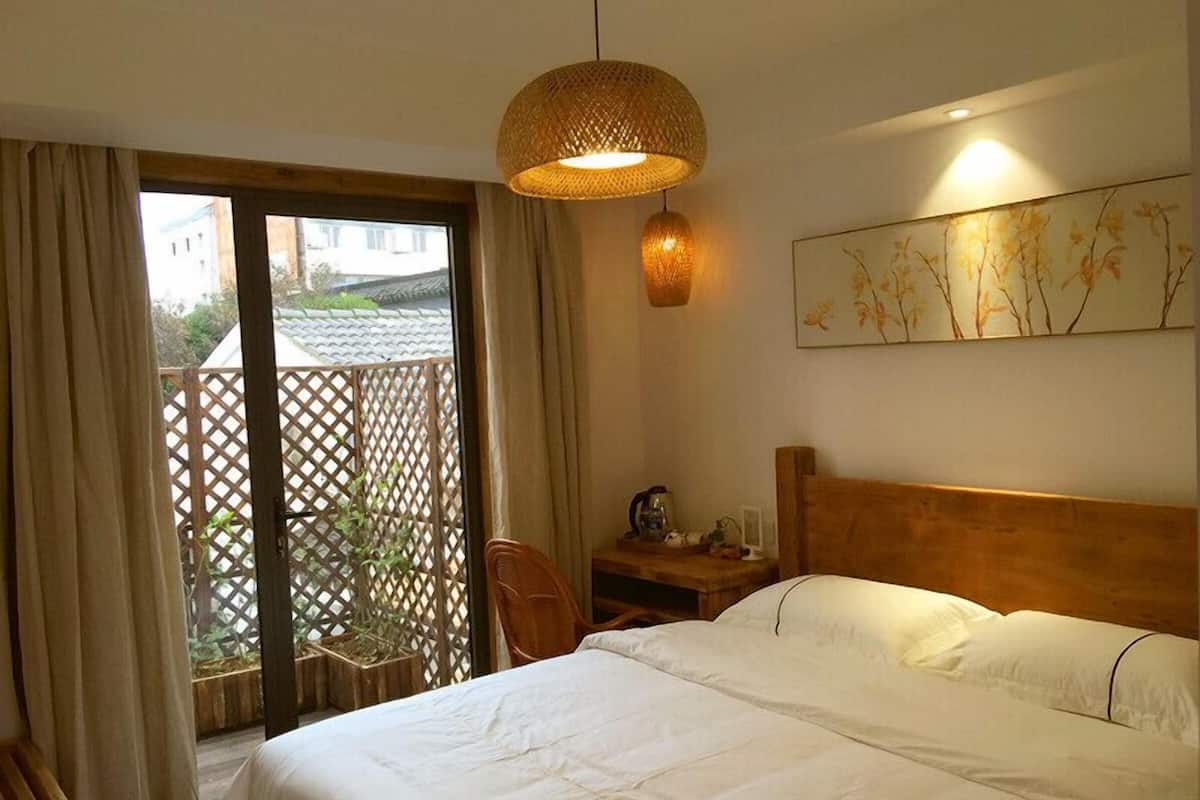 Quarto de hotel com cama, cabeceira de madeira, mesa de cabeceira, abajur e vista exterior por uma porta de correr.