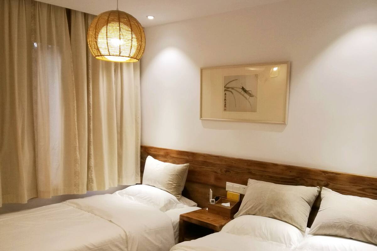 Quarto de hotel com duas camas, cabeceira de madeira, mesa de cabeceira com abajur e um quadro na parede.