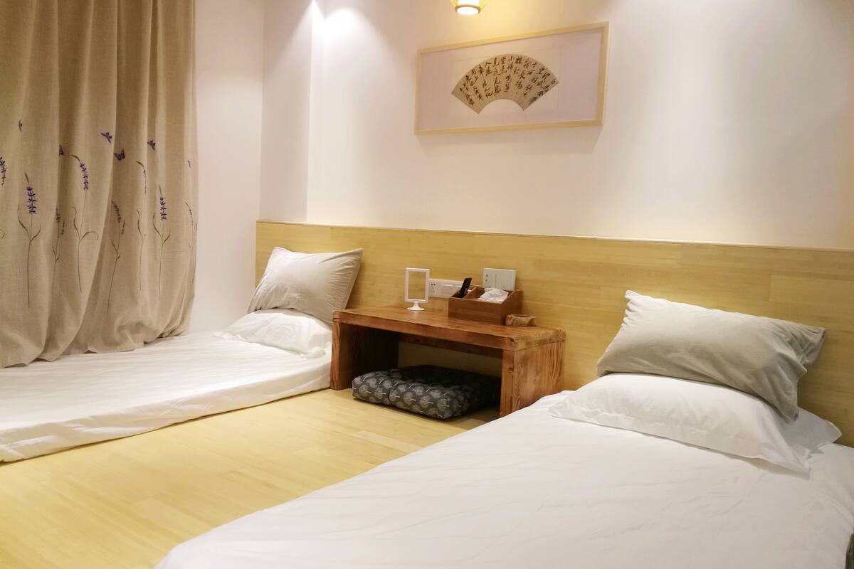 Quarto de hotel com duas camas de solteiro, uma mesa de cabeceira de madeira com abajur, uma obra de arte emoldurada na parede e uma janela com cortinas.