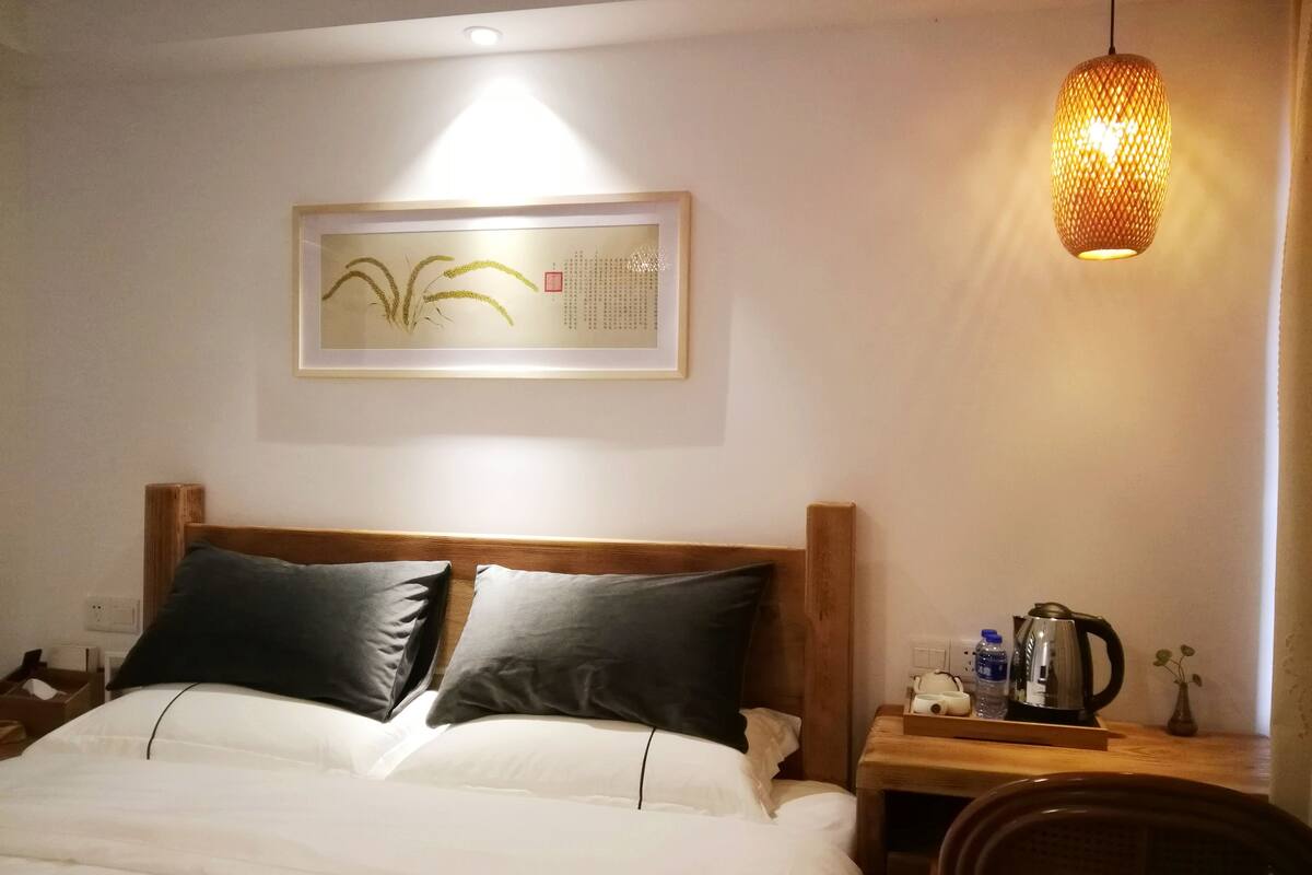 Cama bem arrumada com dois travesseiros, mesa de cabeceira com chaleira e um vaso, e luminária fixada na parede.