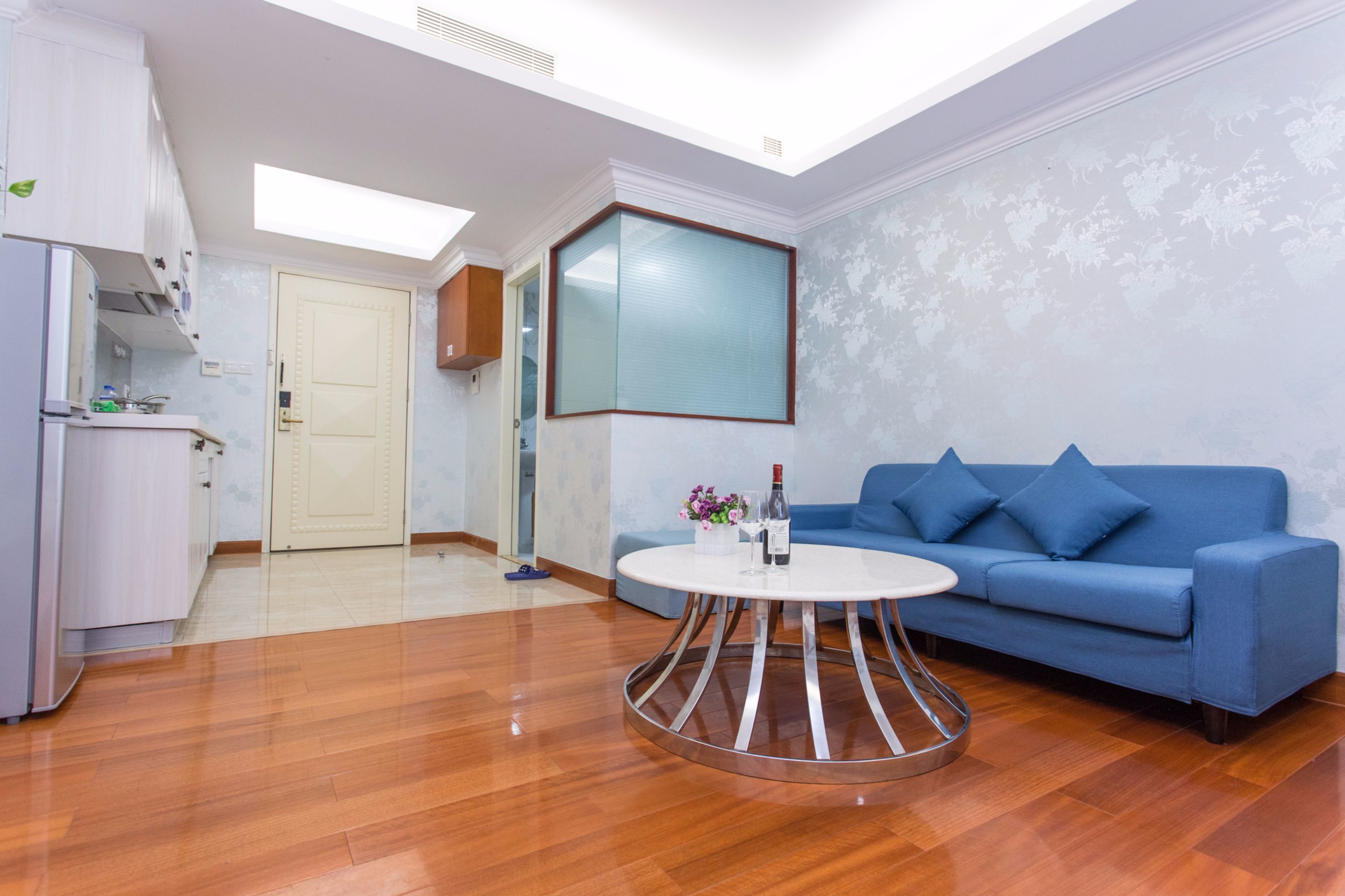 A modern living room with a blue sofa, a round coffee table, and a kitchen area with cabinets and a refrigerator.