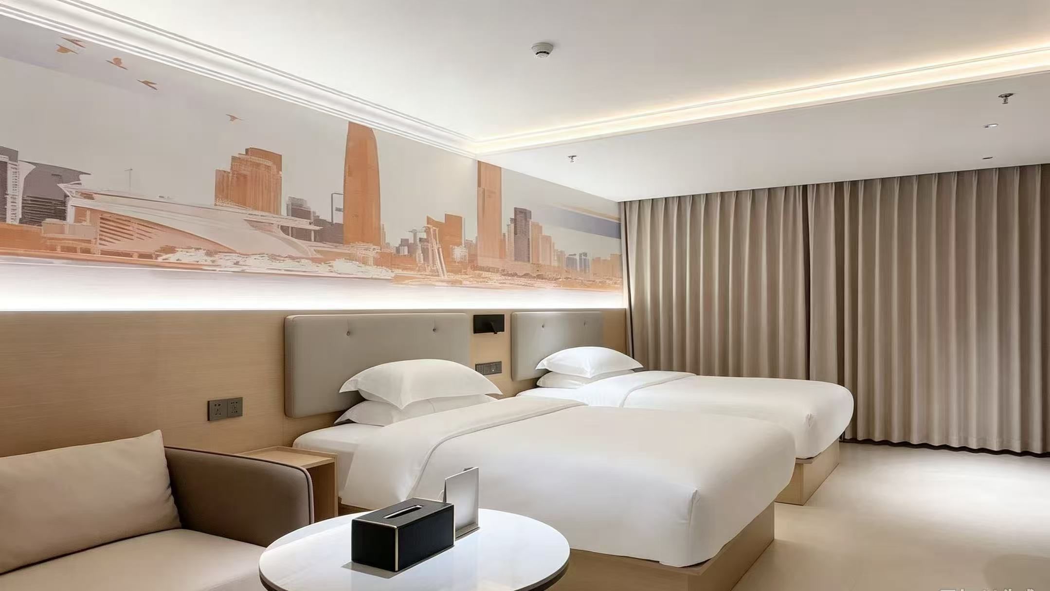 Ein Hotelzimmer mit zwei Betten, einem Sofa, einem kleinen Tisch und einem Wandgemälde mit Stadtansicht.
