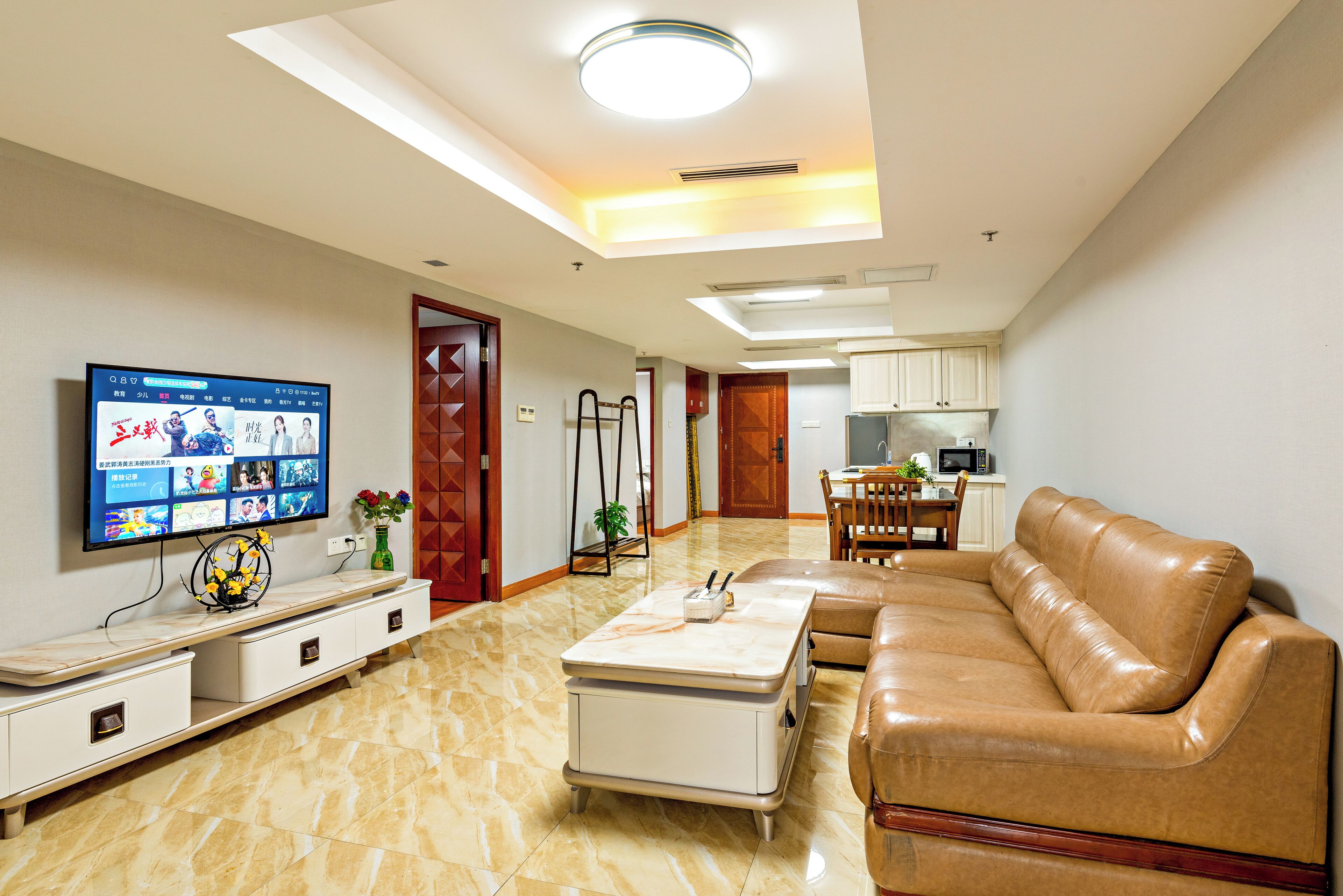 Una sala de estar moderna con un sofá de cuero, un televisor montado en la pared, un comedor con mesa y sillas, y una cocina al fondo.