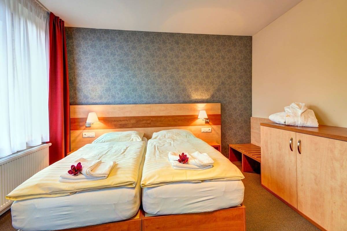 A double bed with white linens, a wooden headboard, bedside lamps, and a wooden cabinet with towels.