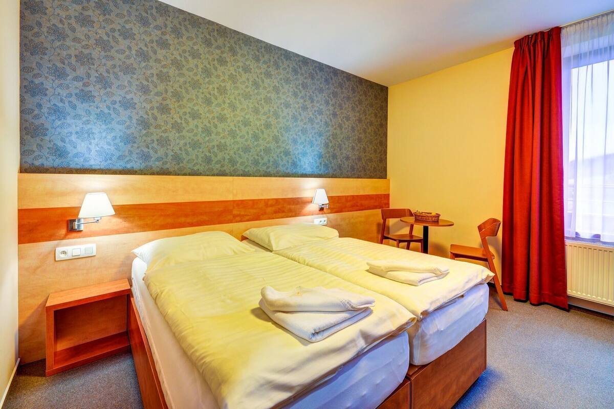 A hotel room with two beds, a small table, a chair, a lamp, and a window with red curtains.