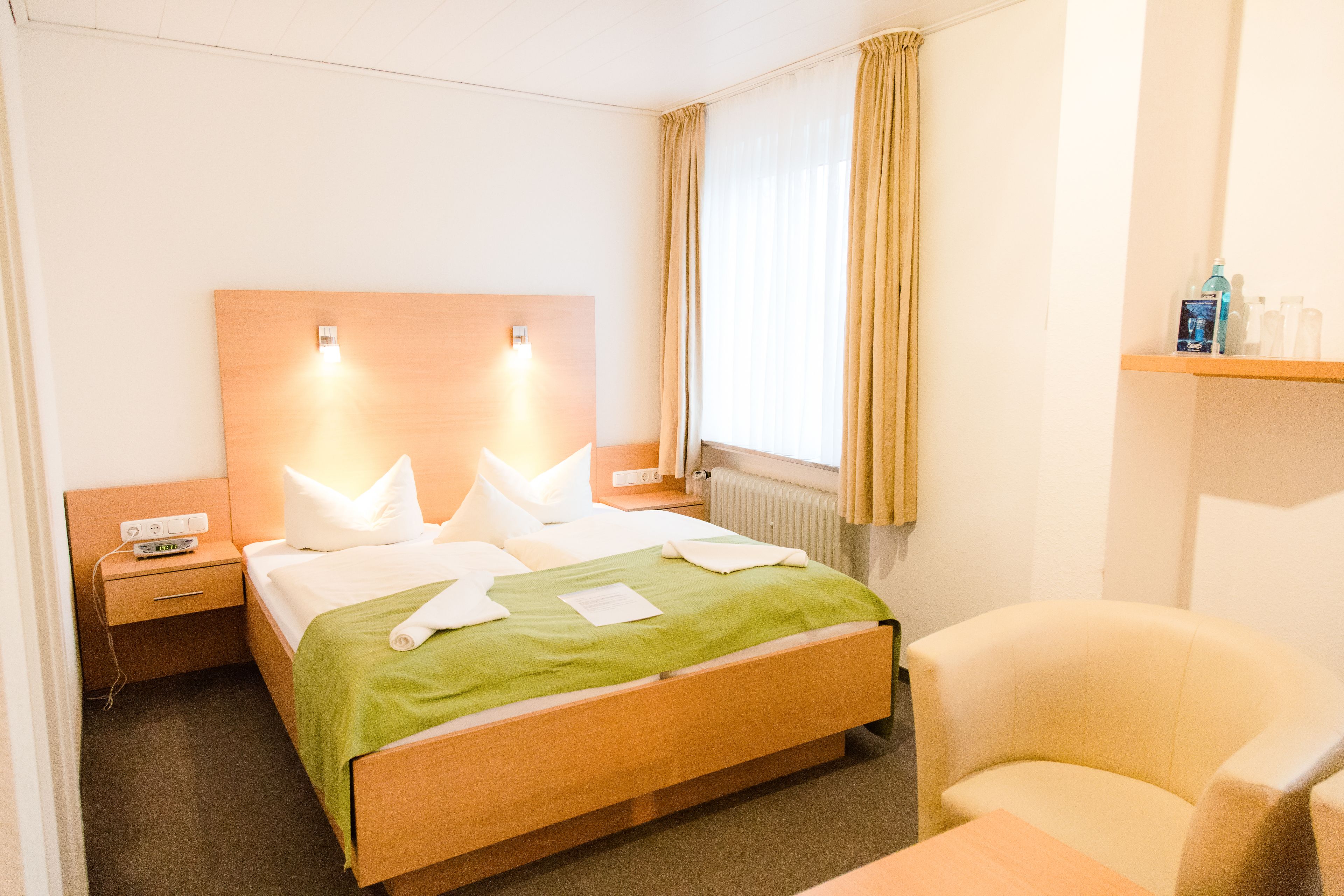 Een hotelkamer met een houten bed, een nachtkastje met een lamp, een stoel en een raam met gordijnen.