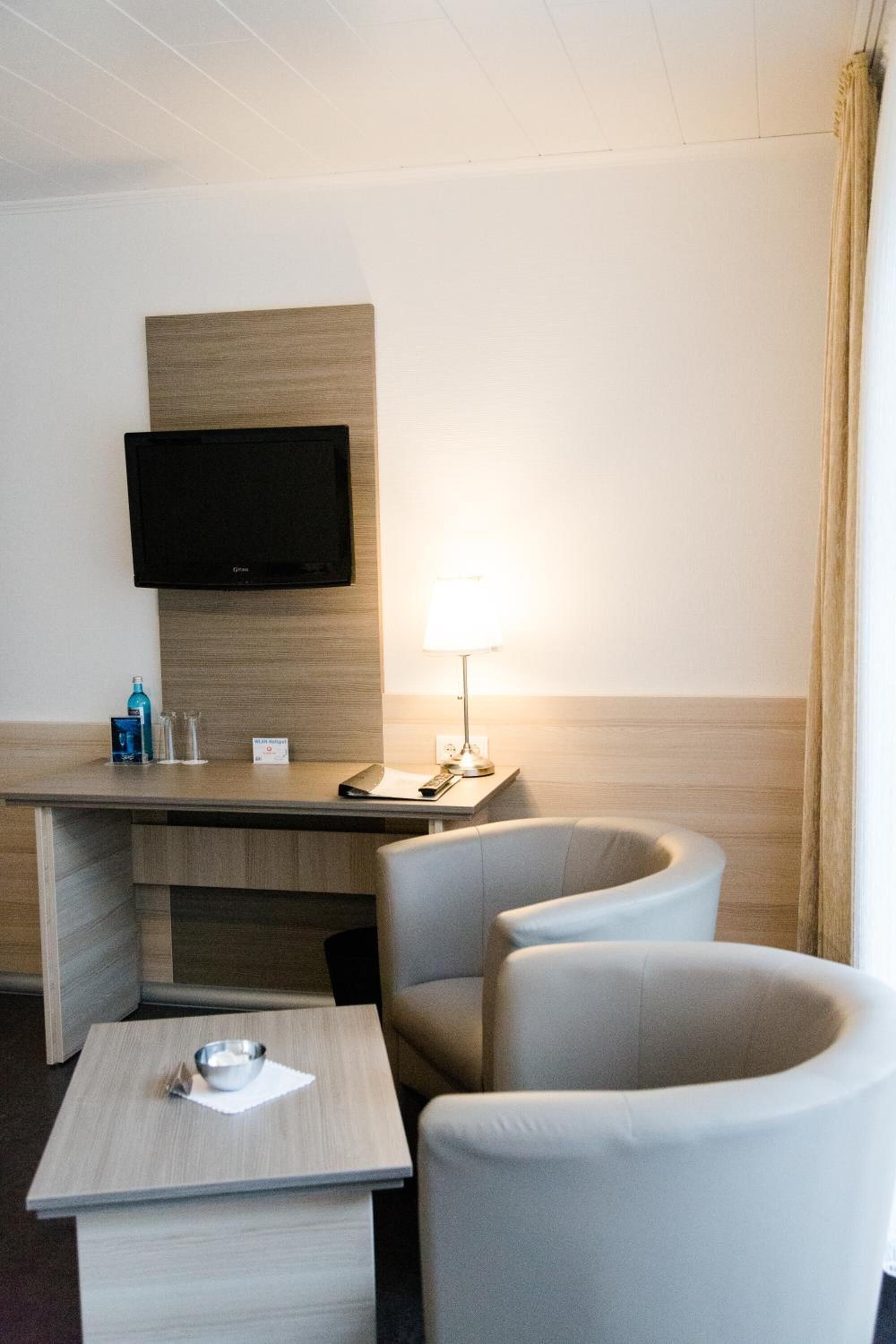 Une chambre d’hôtel équipée d’un téléviseur à écran plat fixé au mur, d’un bureau avec une lampe, de deux fauteuils et d’une petite table sur laquelle se trouve un bol.