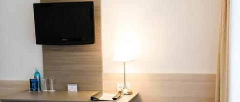Habitación de hotel con televisor de pantalla plana montado en la pared, escritorio con lámpara, dos sillones y una mesita con un cuenco.